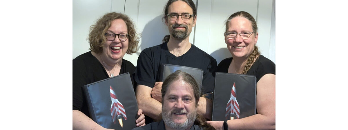 Barbership at DFDF in 2024 - we are all older and greyer, and we hold up our note folders with our Barbership logo: A red and white (polecat) rocket ship.