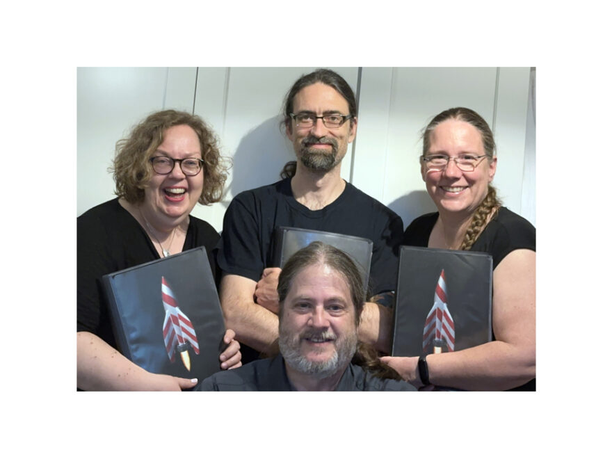 Barbership at DFDF in 2024 - we are all older and greyer, and we hold up our note folders with our Barbership logo: A red and white (polecat) rocket ship.